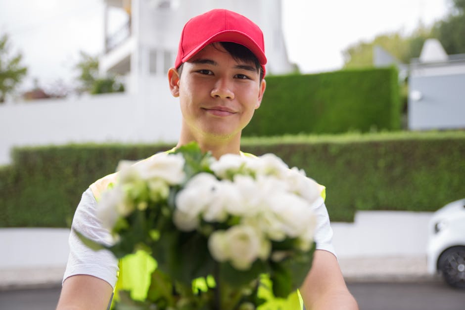 A young male flower delivery courier in a red cap and high-visibility vest is standing outdoors on a residential street, holding a bouquet of fresh, white flowers with green foliage. The bouquet featu