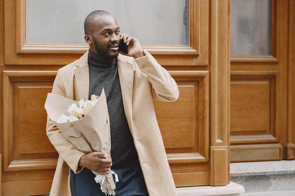 A man with a dark complexion and short black hair stands outdoors in front of a wooden-paneled building, holding a bouquet of fresh white and cream flowers wrapped in brown craft paper. He is wearing 