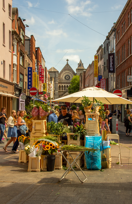 A bustling street scene featuring an outdoor flower stall stocked with vibrant and fresh floral arrangements. The stall displays a variety of flowers including sunflowers, pink peonies, and green foli