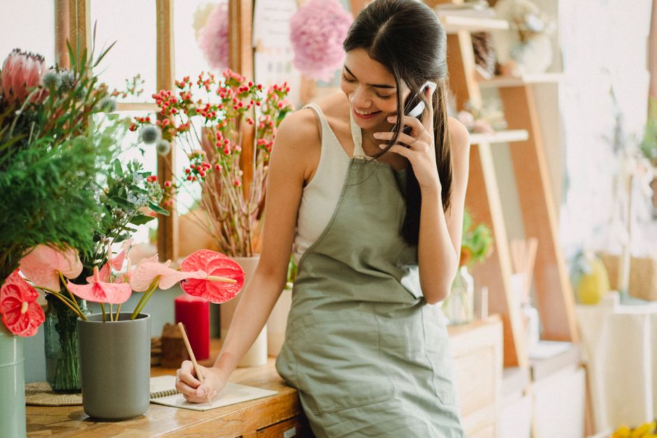 A young female florist with long dark hair, dressed in a white sleeveless top and a light green apron, is smiling while speaking on a black cordless phone. She is writing on a notepad with her left ha