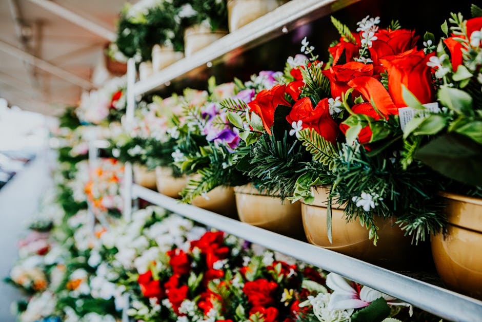 A row of floral arrangements displayed on a metal shelf at a flower shop, featuring vibrant red roses, white and purple blooms, and various green foliage. The flowers are arranged in beige, round pots