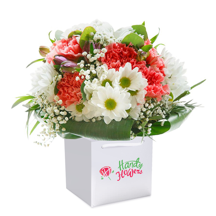 Capturing the refined vibrancy of St James's, this spectacular bouquet presents a rich palette of floral beauty arranged upright in a minimalist, square white gift bag. The mix begins with plush coral and peach-toned carnations, their delicately ruffled petals recalling the exuberant beds of St James's Park in early summer. Interspersed among them, crisp white daisies smile with fresh lime-green centers, offering pops of cheer amid the soft pastels. A light dusting of baby's breath floats gently among the fuller blooms, its petite blossoms bringing an airy quality to the design. In a nod to sophistication, unopened flower buds with hints of purple and pink peek discreetly from the left side, adding depth and echoing the subtle shades seen near the Royal Academy's floral installations. The whole arrangement is enveloped by generous, dark glossy foliage, which gracefully fans out to create a striking contrast and structural harmony. Ready for delivery throughout St James's, this bouquet sits snugly in its modern white gift bag, sturdy handles poised for easy carrying along Pall Mall or Piccadilly. The composition speaks to the pride, freshness, and artistic flair of local florists-making it ideal for life's special celebrations or simply brightening a stylish London flat.