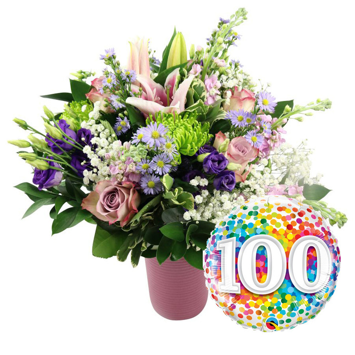 On a bright white background, a joyful centenary scene unfolds: a generous, pastel-toned bouquet in a ribbed matte pink vase is accompanied by a festive foil balloon bearing the number 100. The bouquet's shape is rounded and full, suggesting considered craftsmanship from a local St James's florist familiar with elegant West End tastes. At the top, several light pink lilies open outward, their star-shaped petals slightly curved, each bloom revealing fine freckling near the centre and pronounced stamens that add sculptural detail. Some lily buds remain closed, slender and tall, offering vertical interest and a sense of time passing gently. Around these prominent flowers cluster soft pink roses and deeper dusty mauve blooms, their petals layered in tight spirals and looser curls, evoking both luxury and tenderness. Through this romantic mix, stems of deep purple lisianthus wind delicately, and airy branches of small lavender asters, with their yellow centres, look as if they have been gathered from a sunlit border near St James's Park. Lime-green button chrysanthemums are tucked between the other blooms, their compact, rounded heads injecting freshness and energy. Threads of white baby's breath drift through the arrangement, creating a light, cloud-like effect that balances the richer colours. Framing everything is a backdrop of glossy dark green foliage, enhancing the sense of garden freshness. To the lower right of the flowers, a round foil balloon shines with an all-over pattern of tiny confetti dots in vivid red, orange, yellow, green, blue, and purple. At its centre, large white numerals 100 appear bold and slightly raised, thanks to a gentle shadow effect. Together, bouquet and balloon convey a mood of joyful respect and enduring achievement, suitable for a 100th birthday tea at a St James's club, a centenary celebration in an office near The Mall, or a family gathering in a nearby flat.