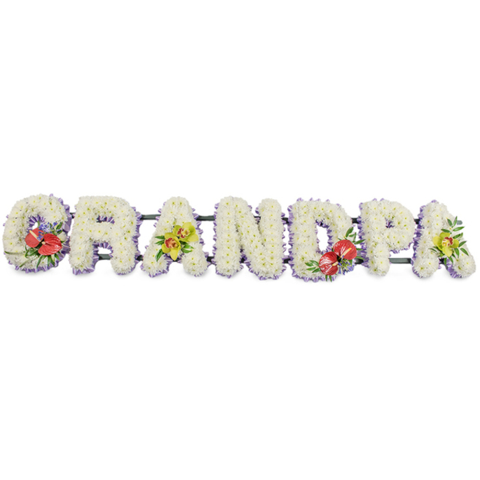 In this image, the word GRANDPA is lovingly spelled out in large, upright block letters created entirely from fresh flowers, arranged in a straight horizontal line on a white, uncluttered background that emphasises every detail. Each letter is crafted from tightly packed white chrysanthemums, their compact blooms forming a plush, even surface with small, petal-rich domes that evoke softness and calm. Around the bottom of the letters runs a pale lavender ribbon trim, offering a gentle hint of colour that delicately outlines the word without overpowering its simple beauty, much like the understated charm of the streets around St James's Park and The Mall. The letters are affixed to dark green structural bars that quietly support the design, giving it the strength needed for a funeral cortege or display beside a coffin. Splashes of richer colour are placed with intention: the first letter G and the D closer to the centre are embellished with vibrant clusters of red anthuriums, their glossy, heart-shaped forms and bright spadices standing out vividly against the white, surrounded by lush greenery and small purple filler blooms. The first and last A hold elegant groupings of yellow-green orchids, their refined, star-like blooms and arching stems adding a contemporary grace that feels fitting for a service in the refined St James's area. This floral GRANDPA tribute carries a message of deep respect and affection for a grandfather, suggesting years of shared moments and family gatherings now distilled into one serene, beautifully composed farewell.